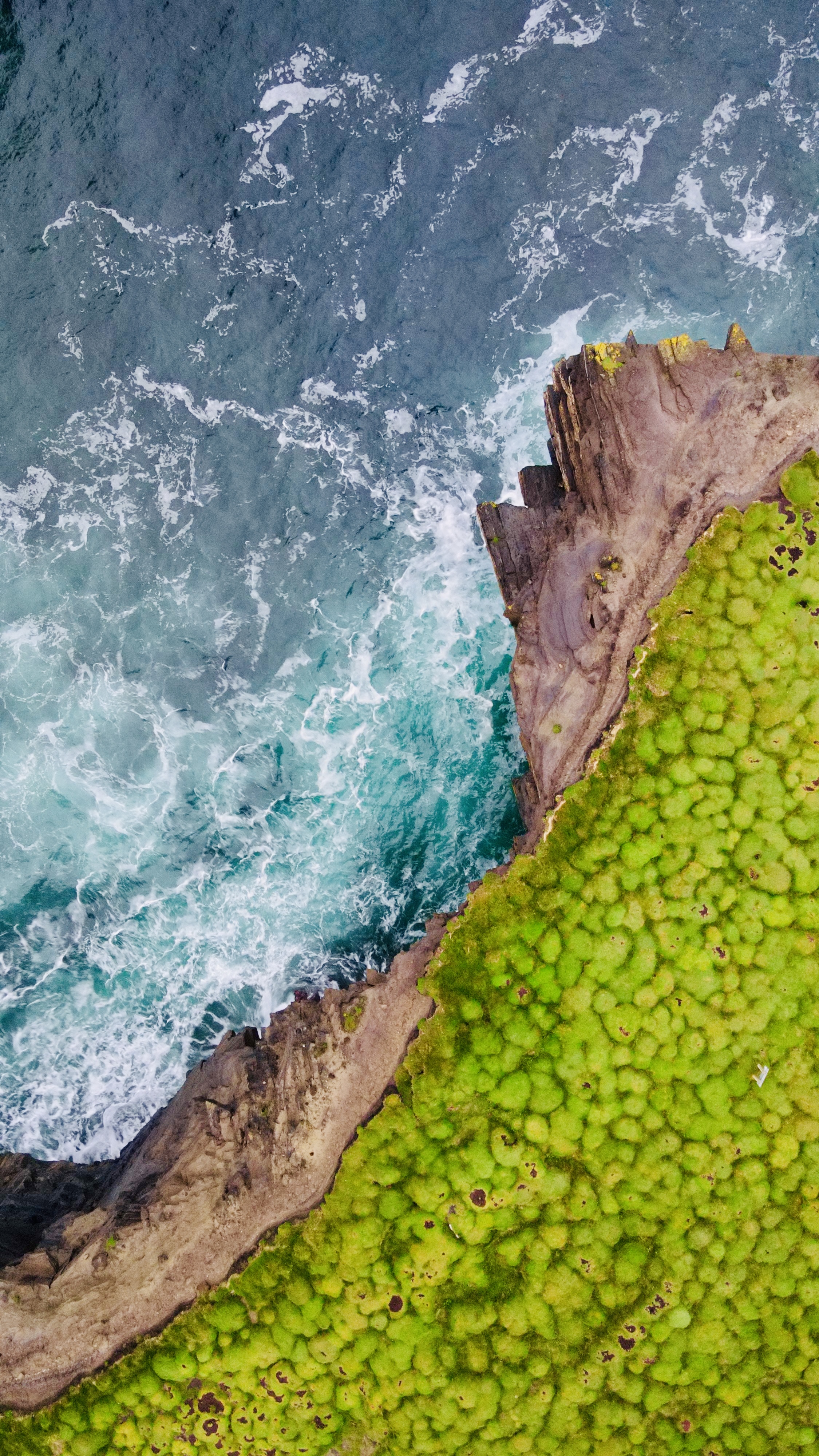 Aerial view of ocean waves meeting a moss-covered cliff